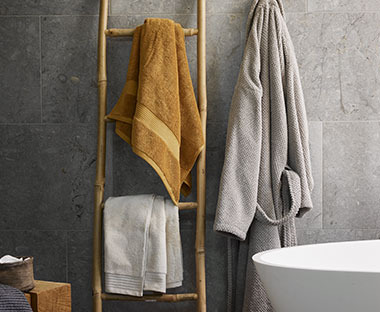 Ein hellgraues und ein senfgelbes Handtuch hängen an einem Leiterregal, daneben hängt ein Bademantel