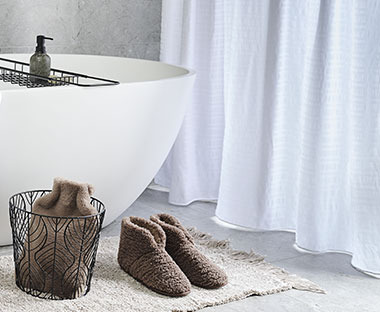 Tenda da doccia bianca affianco a vasca da bagno, pantofole marroni e borsa dell'acqua calda in un cesto su un tappetino da bgno