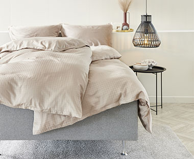 Hellbeige Bettwäsche aus Satin auf einem grauen Bett, daneben schwarzer Nachttisch