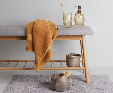 Panca in legno e stoffa su di essa accessori da bano e asciugamano e sul pavimento tappeto beige e contenitore in vimini