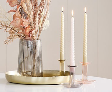 Portacandele colorate con candele affiancate da vassoio dorato e vaso grigio trasparente e fiori artificiali