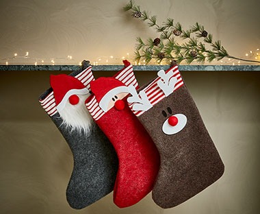 Tre calze di Natale di colore grigio, rosso e marrone e diverse figure appese al camino vicino le luci