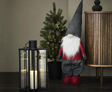 Lutin de Noël à côté d'une lanterne noire, d'un arbre de Noël et d'une armoire en bois.