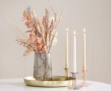Farbige Kerzenständern mit Kerzen auf goldenem Dekotablett neben einer grauen Vase mit Kunstblumen