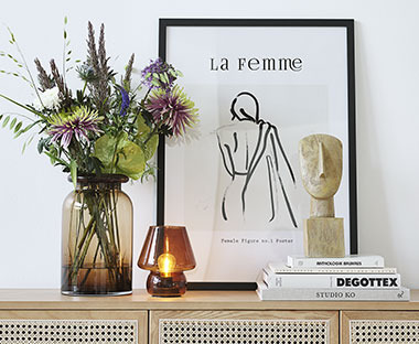 Bild mit schwarzem Rahmen, Vase mit Blumen, kleine Dekolampe und eine Skulptur auf einem Sideboard