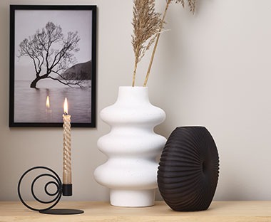 Weisse Vase mit weichen Rundungen, daneben ein schwarzer Kerzenständer und eine Vase in Muschel-Form