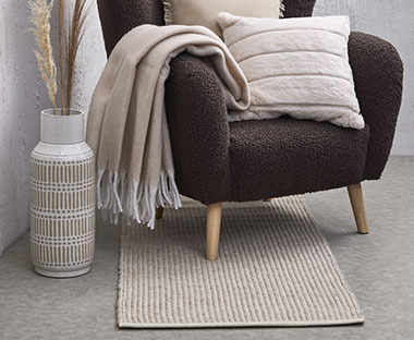 Heller, eckiger Teppich, darauf ein dunkelgrauer Sessel mit Zierkissen und Decken dekoriert, daneben ist eine weiss-beige Vase aufgestellt