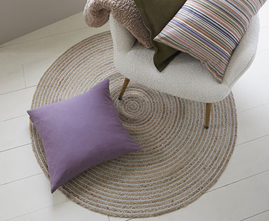 Runder Teppich aus Jute, darauf ein lila Zierkissen und ein weisser Sessel