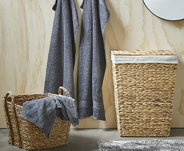 Eckiger Wäschekorb aus Stroh und ein kleiner Korb davor, zwei blaue Handtücher an einer Wand