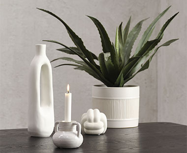 Table noire avec une plante artificielle dans un cache-pot blanc, flanquée d'un bougeoir, d'une sculpture et d'un vase tous blancs