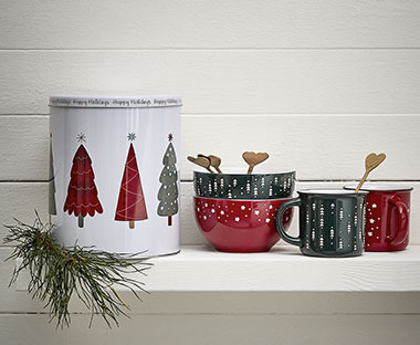 Boîte à biscuits blanche avec motifs de sapins de Noël à côté de tasses et bols rouges et verts avec des cuillères à café dorées