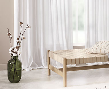 Des rideaux blancs sur mesure dans un salon flanqué d'un banc en bois et d'un vase vert avec de fausses fleurs