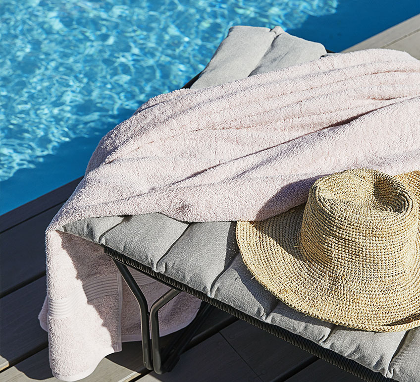 Sonnenliege mit einer grauen Gartenauflage, Badetuch und Strohhut an einem Pool