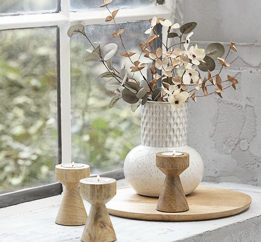 Beige-weisse Vase geprägt mit einem Muster auf einem Holztablett auf einem Fenstersims, Kunstblumen und Teelichhalter aus Holz