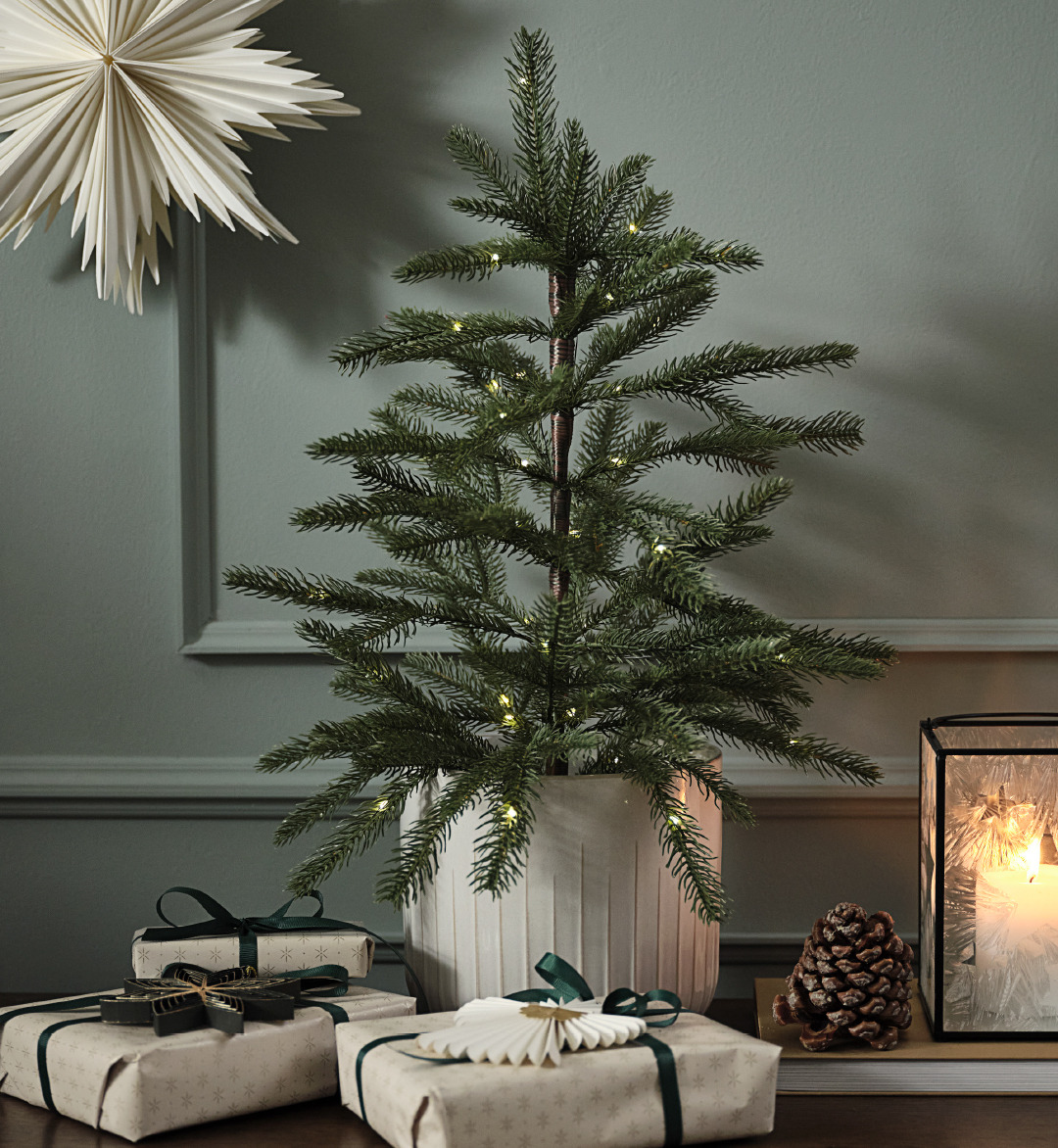 Künstlicher Weihnachtsbaum im Topf mit Lichtern und einem Papierstern an der Wand