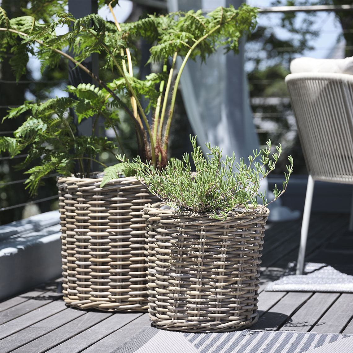 Fioriere SEGLA Due cestini porta-piante in rattan naturale non trattato posizionati su un balcone, con piante rigogliose che aggiungono un tocco di verde e freschezza allo spazio esterno