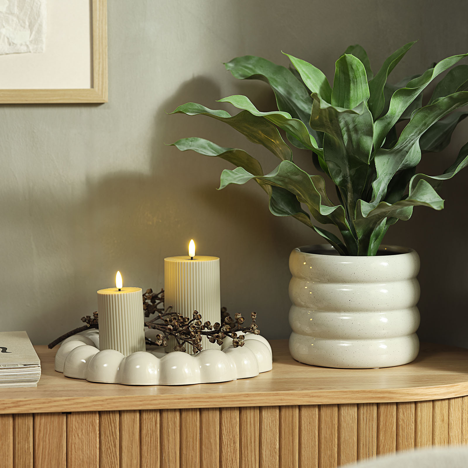2 bougies allumées sur un plateau décoratif blanc chaud avec des bords "bobble" à côté d'un cache-pot blanc chaud