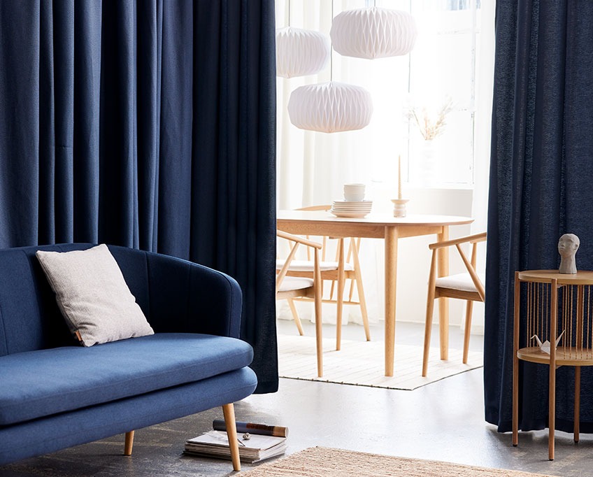 YMSEN curtain behind GISTRUP sofa and EGELEV end table with MARSTRAND dining table, ARNBORG dining chairs and VALDER lamps in the background Blue curtain behind a sofa separates a living area from a dining area