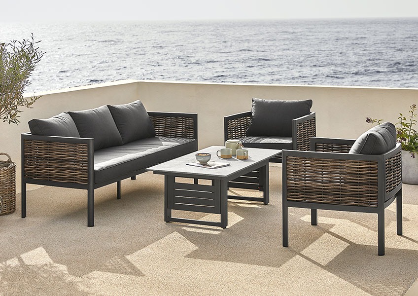 Salon en rotin marron et coussins noirs confortables sur un grand balcon avec vue sur la mer.