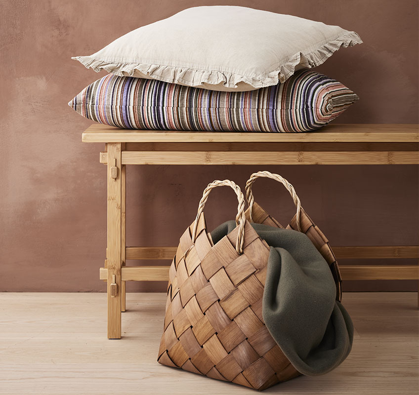 Zierkissen BANKSIA und GULDBLOMME, Korb ABEL und Fleecedecke DAGLILJE Dekorative Zierkissen auf einer Holzbank und Korb mit einer Wohndecke darin