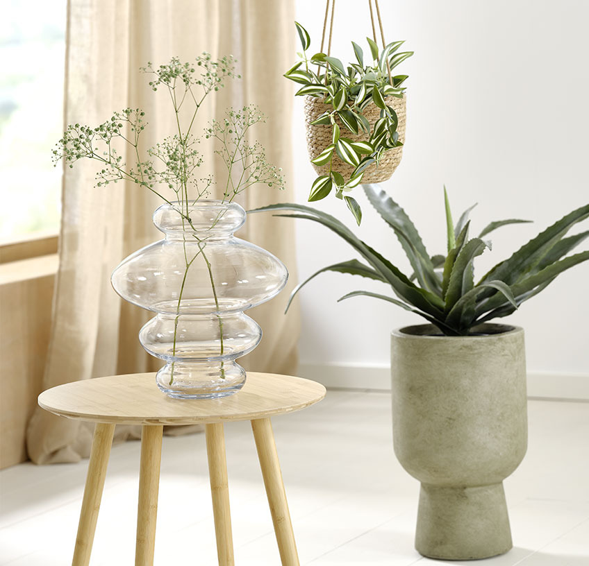 Vase aus Glas auf einem Beistelltisch, Hängepflanzentopf und grüner Pflanzentopf mit Kunstpflanzen