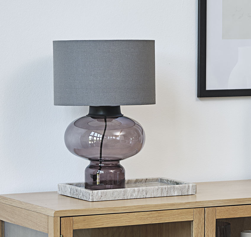 Tischlampe mit Glasfuss und grauem Lampenschirm auf einem Marmortablett