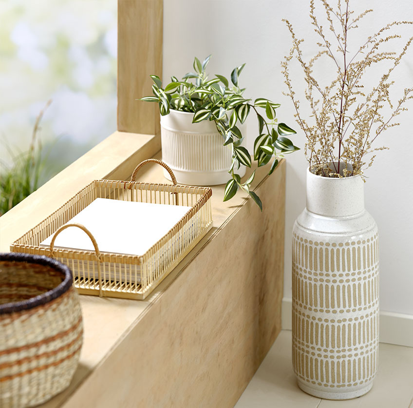 Grosse Vase neben einer Fensterbank, darauf ein Tablett aus Bambus und weisser Pflanzentopf mit Kunstpflanze darin