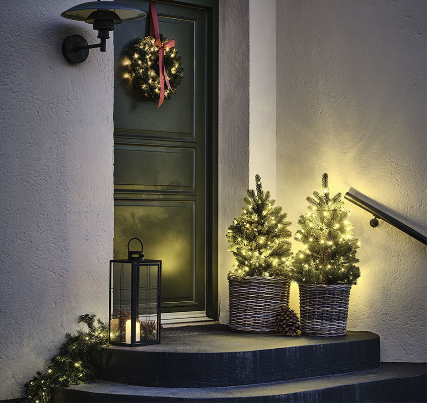 Zwei Kunstbäume mit Lichterketten vor der Haustür und Weihnachtskranz mit roter Schleife